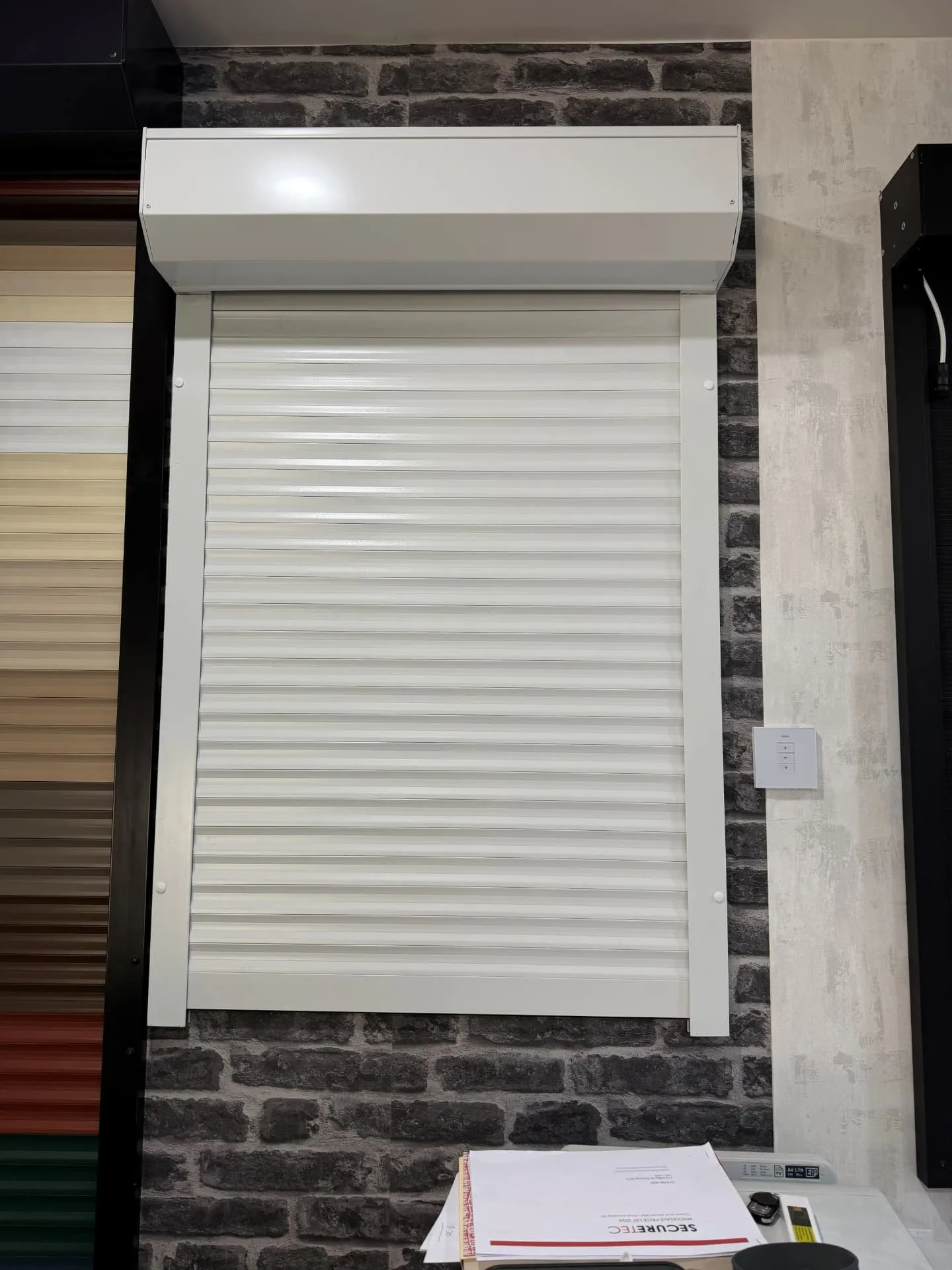 Closed white roller shutter and headbox mounted on a brick-look display wall in the Securetec showroom