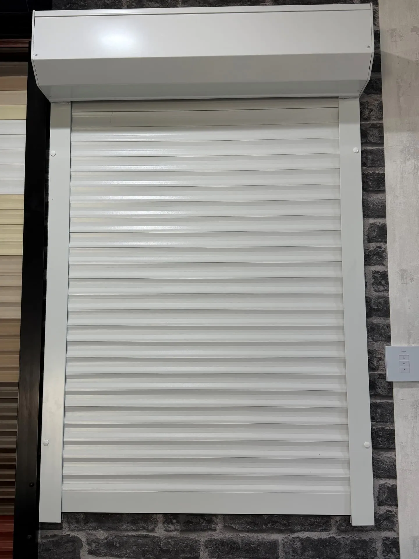White roller shutter with matching headbox and guides on a Securetec showroom display wall, with a wall switch beside it