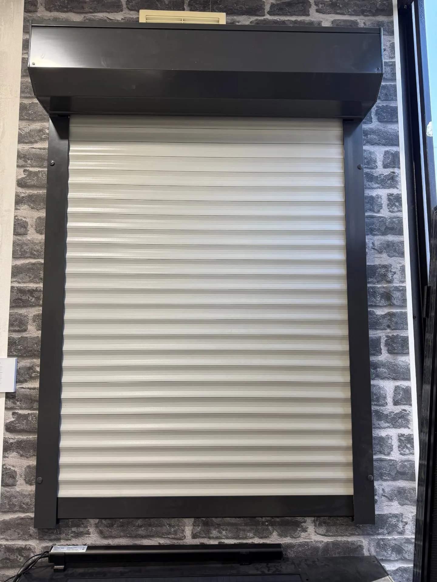 Two-tone showroom roller shutter with light grey slats and a charcoal headbox, guides and bottom bar