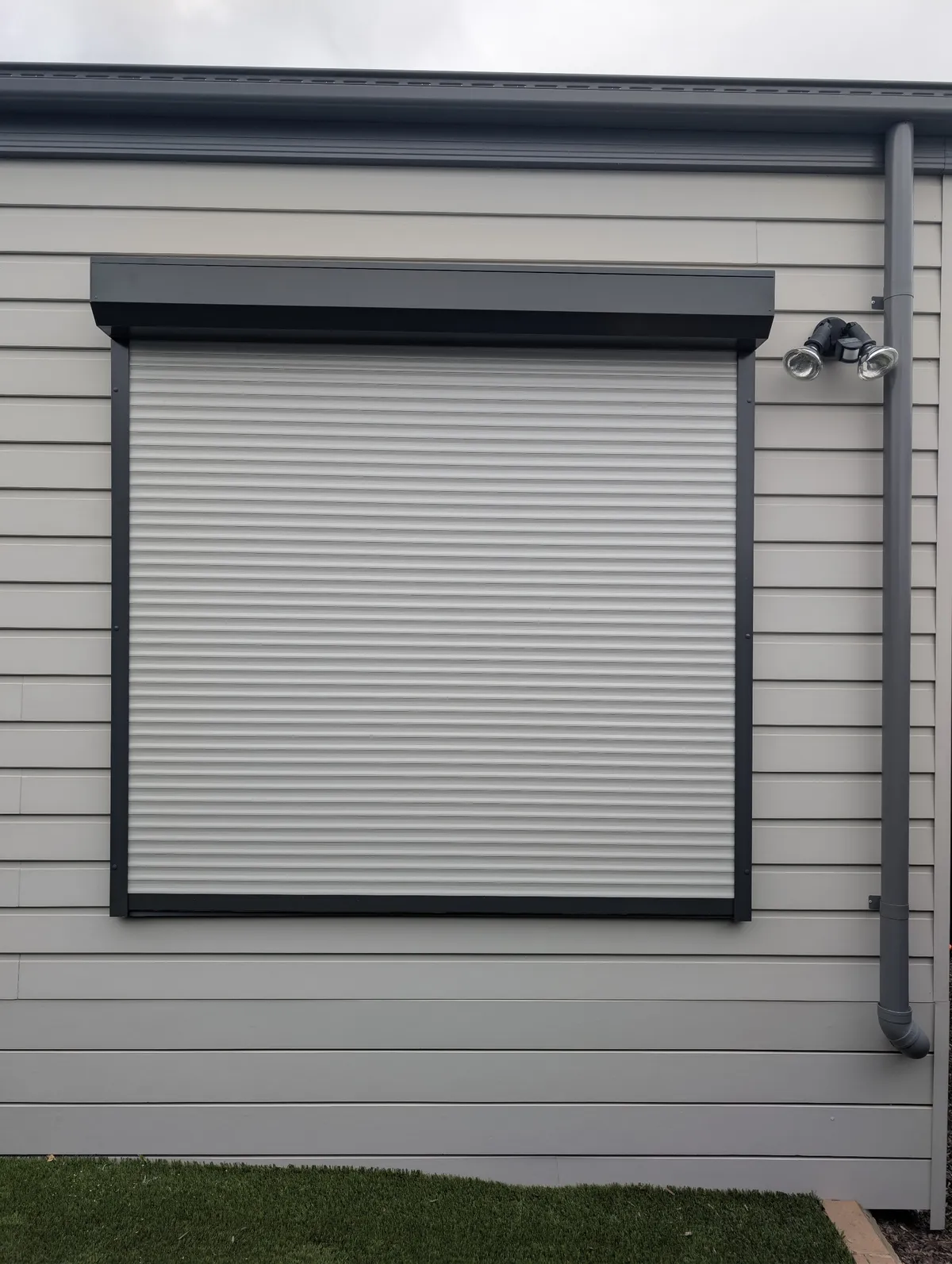 Roller shutter with a dark headbox and guides over a window on a grey weatherboard home