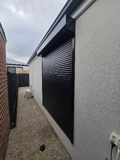 Black roller shutter closed over a tall window on a rendered home beside a side path